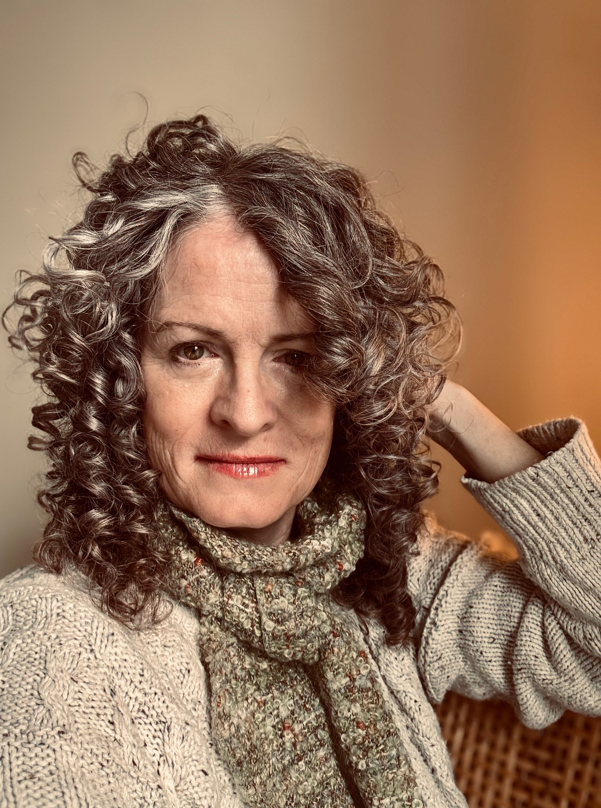 middle aged woman with curly salt and pepper hair looks at the camera. She wears neutral warm colours.